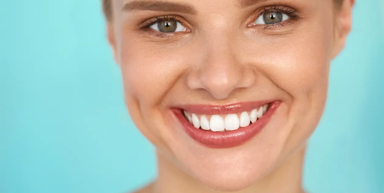Portrait d'une femme souriante aux yeux verts et aux dents blanches parfaites, posant devant un fond bleu clair.