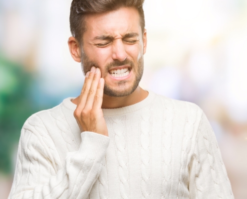 Douleur dentaire intense nécessitant une urgence Un homme en pull blanc tient sa joue avec une expression de douleur intense, indiquant un mal de dents.